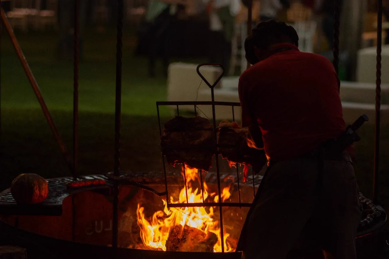 Parrillero cocinando cortes de carne sobre el fuego de un fogón durante un evento nocturno en Las Cortaderas
