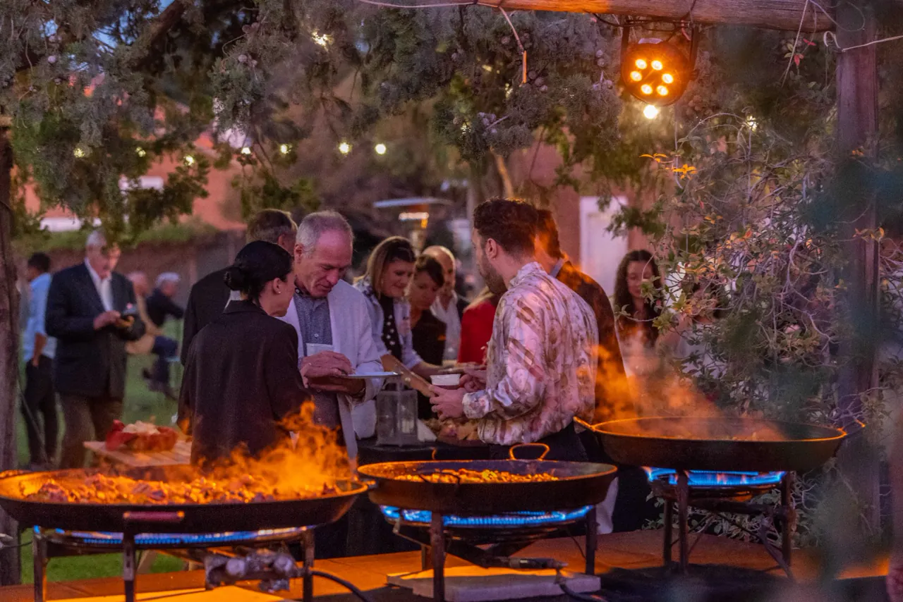 Discos de paella al fuego rodeados de invitados durante un evento al atardecer