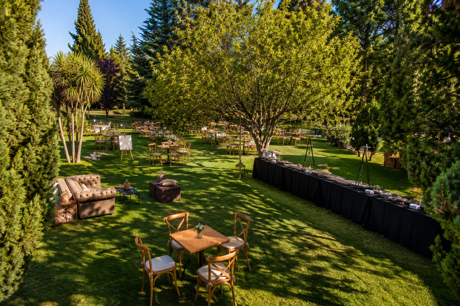 Parque del predio montado de día para un evento, con mesas de madera, livings y árboles bajo el sol