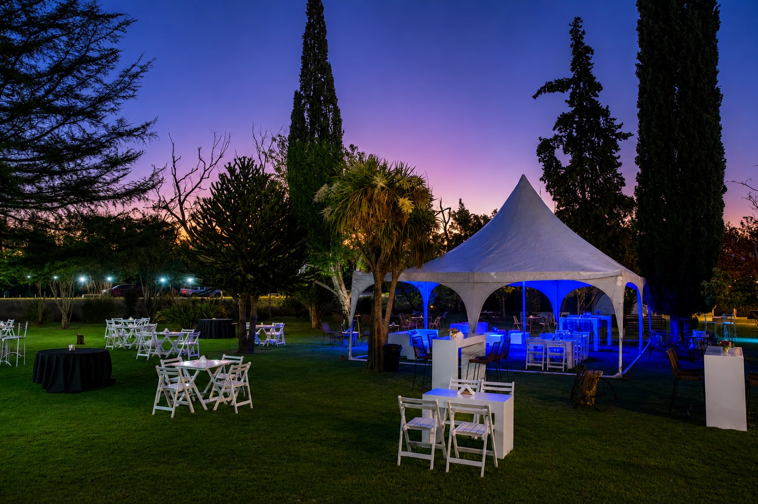 Atardecer violeta sobre el predio, con la carpa iluminada en azul y mesas vestidas en el parque
