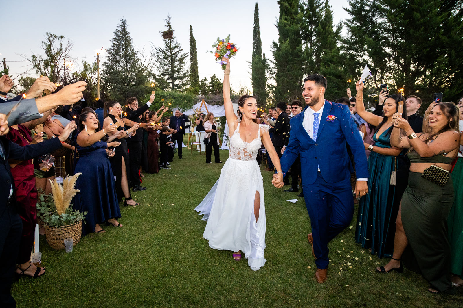 Los novios caminan tomados de la mano entre los invitados que encienden bengalas al atardecer; la novia levanta el ramo